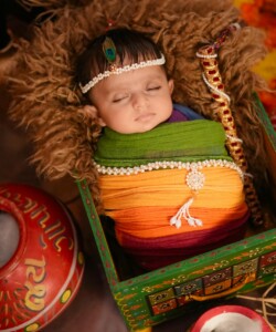 A sleeping baby wrapped in multicolored fabric, adorned with traditional jewelry, surrounded by festive Indian elements.
