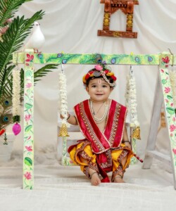 Adorable baby in vibrant traditional clothing sitting on a decorated swing with doves and decorative items.